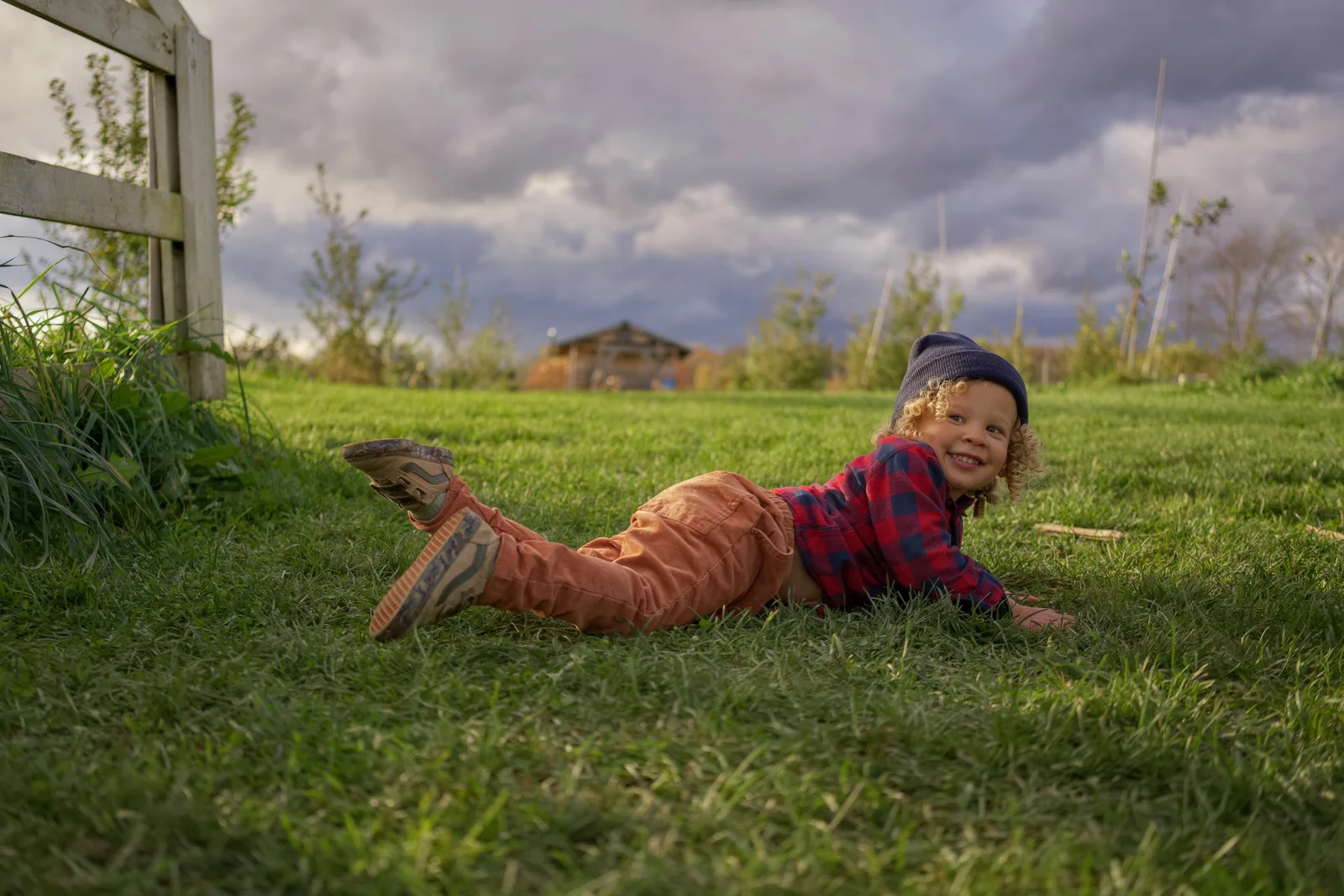 Child in red plaid shirt, orange pants, and gray beanie lying on grass near wooden fence