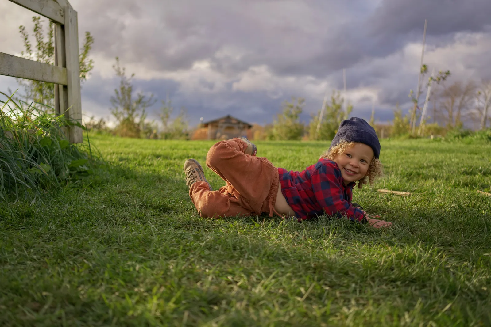 Child in red plaid shirt, orange pants, and gray beanie lying on grass with legs raised