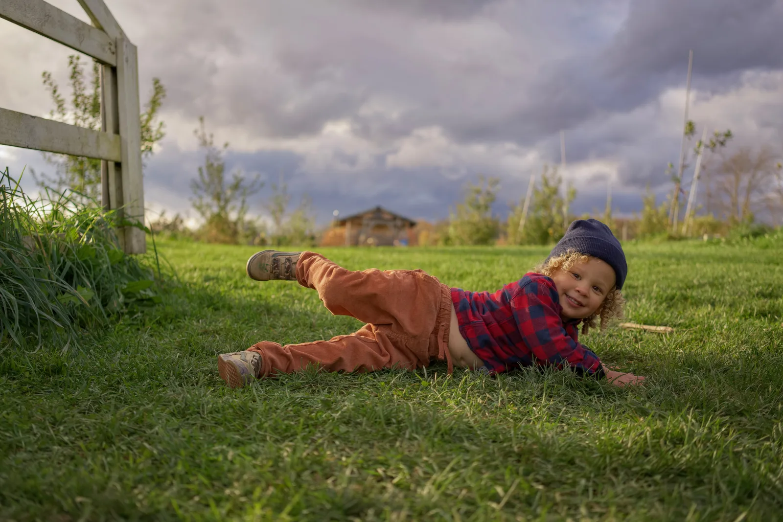 Child in red plaid shirt, orange pants, and gray beanie lying on grass with one foot up