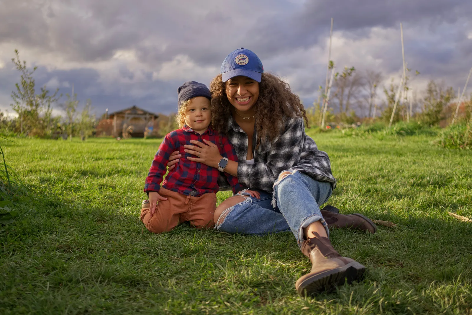 Two people in plaid shirts and caps sitting on grass with barn and cloudy sky behind