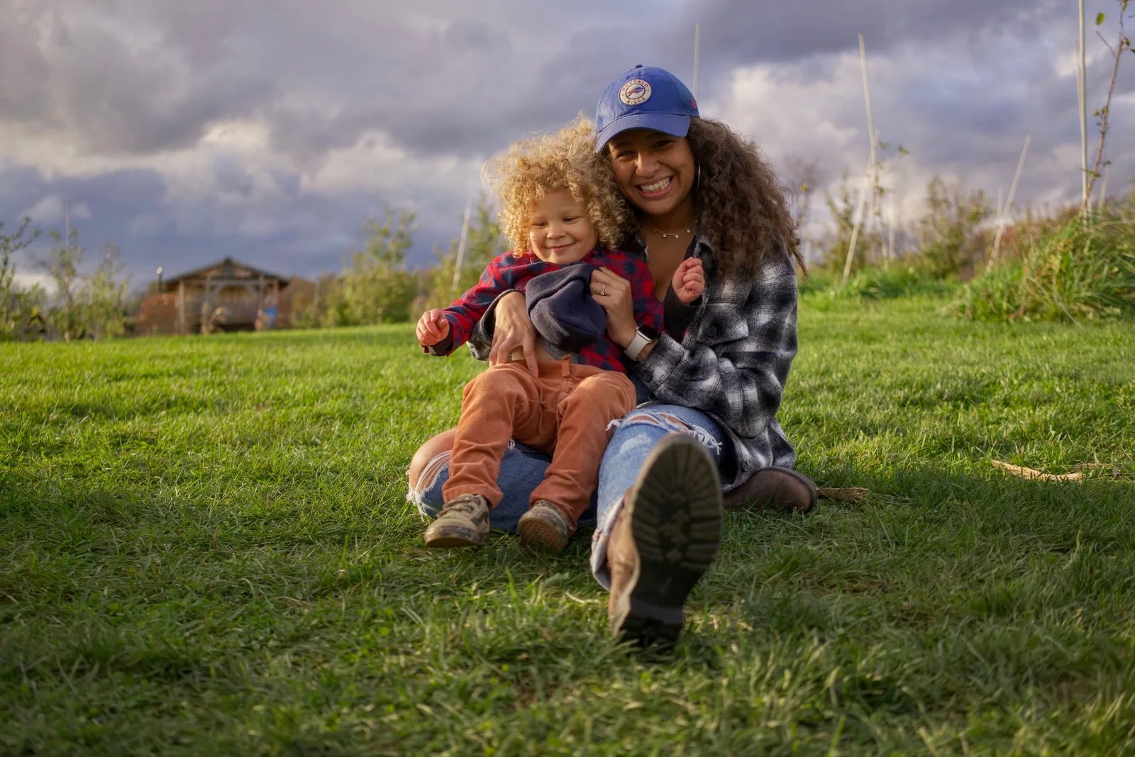 Two people sitting on grassy hill, one in blue cap and plaid shirt holding child in red plaid shirt
