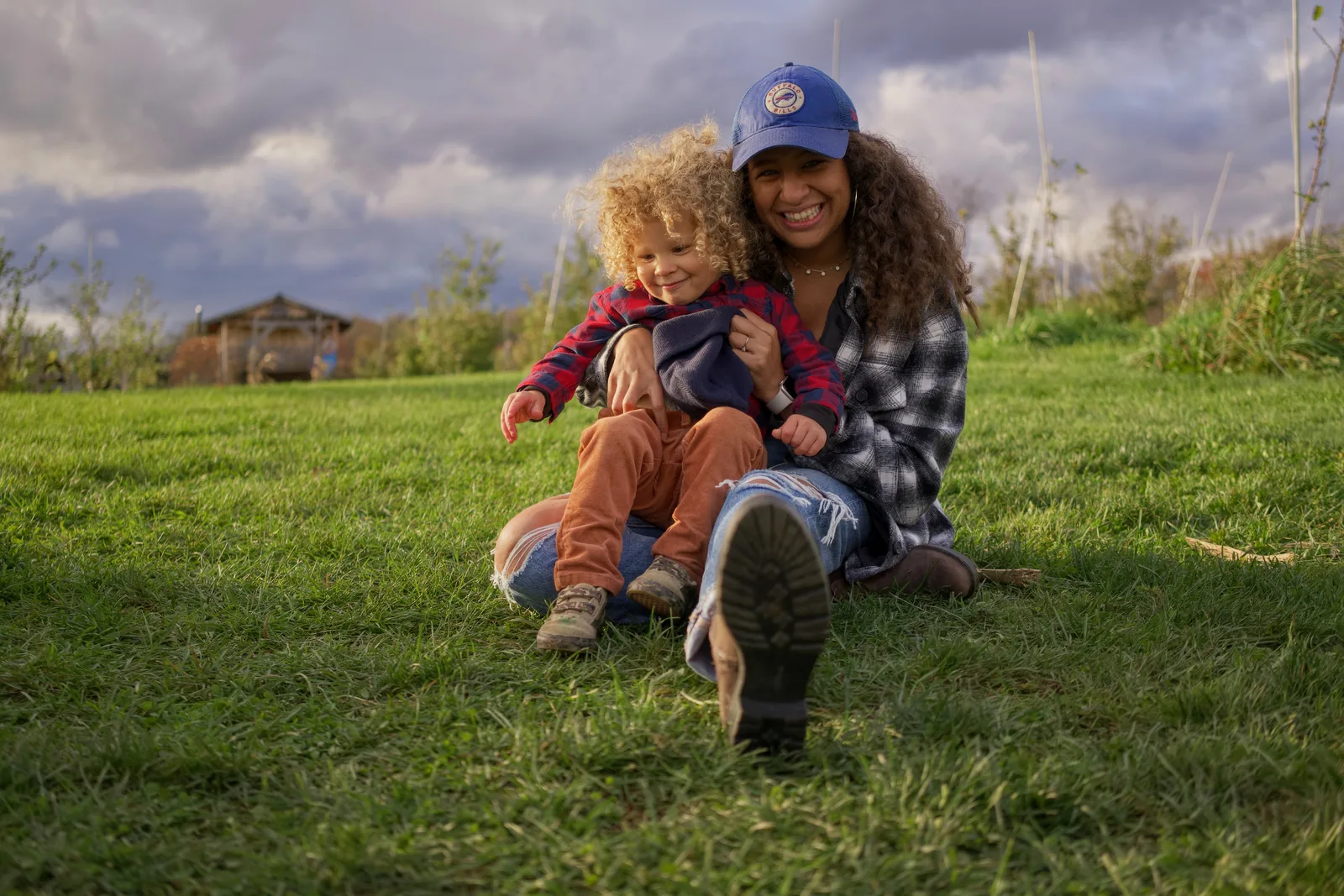 Two people sitting on grassy hill, one in blue cap and plaid shirt holding child in red plaid