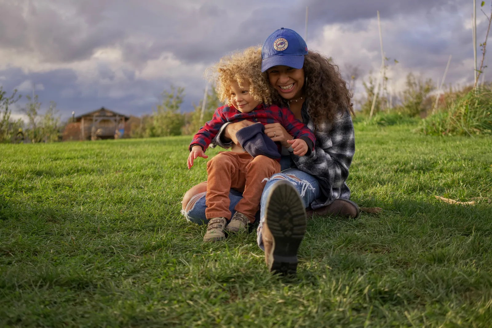 Two people on grassy hill, one in blue cap and plaid shirt holding child in red plaid shirt