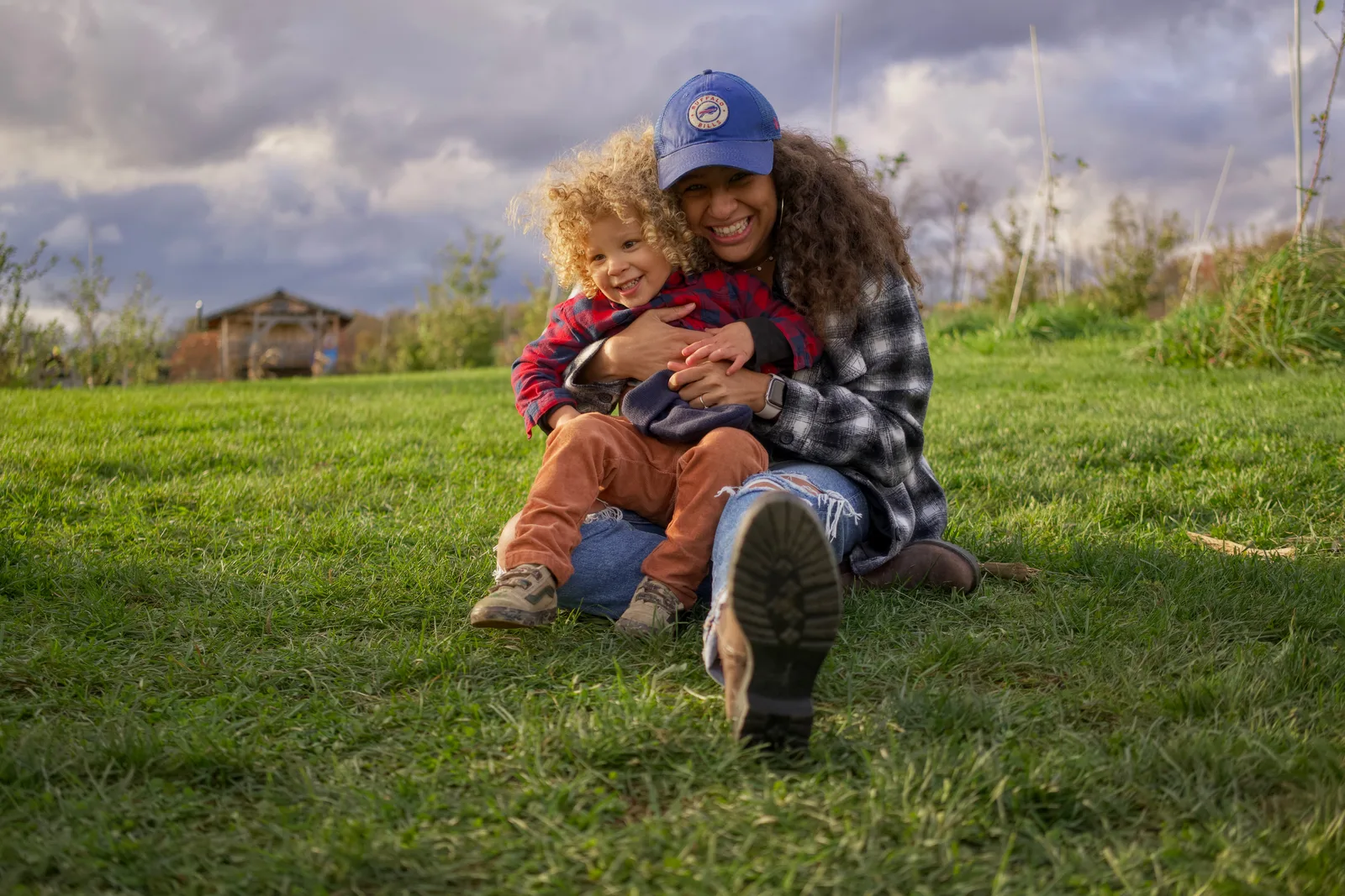 Two people on grassy hill, one in blue cap and plaid shirt embracing child in red plaid