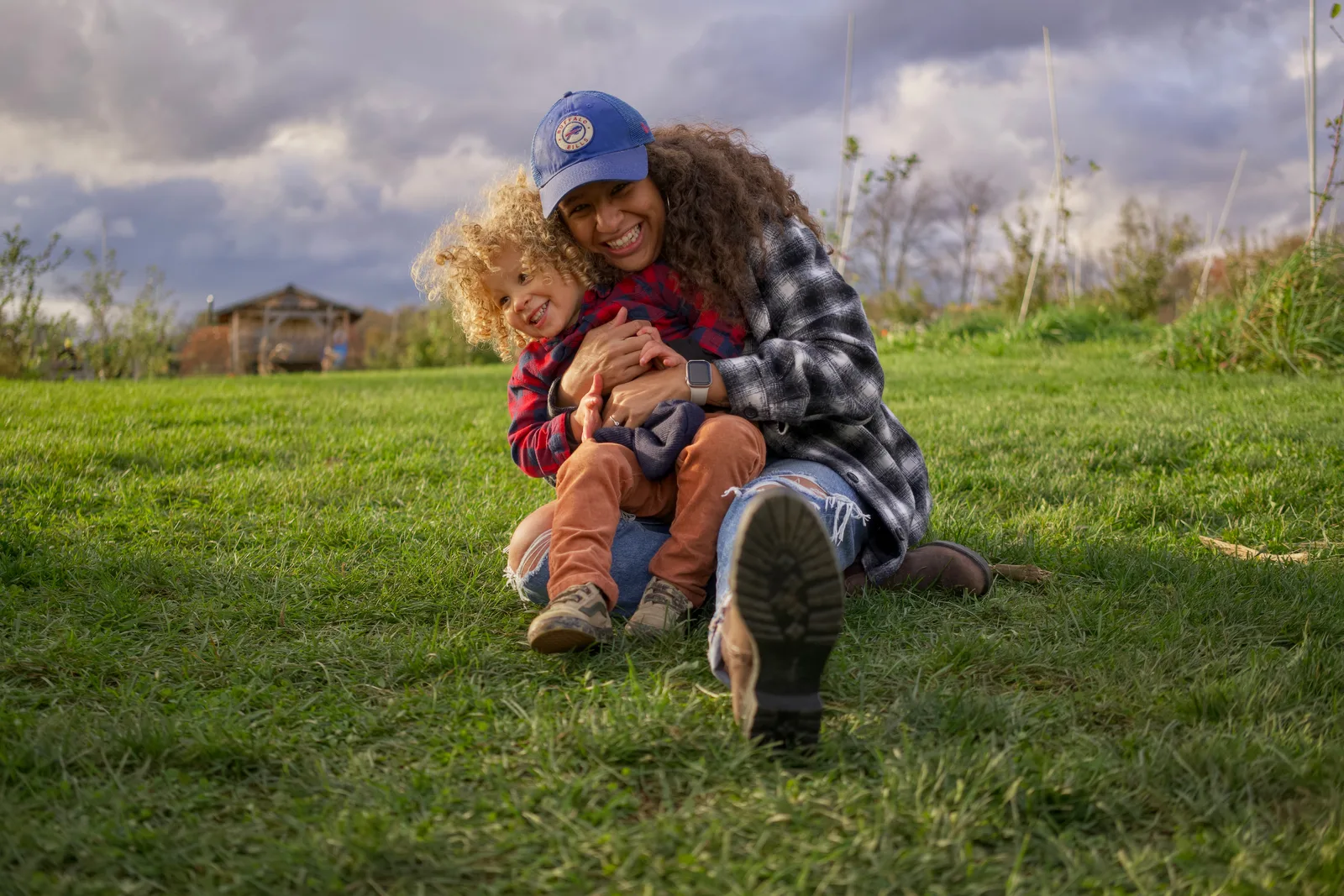 Two people on grassy hill, one in blue cap and plaid shirt embracing child in red plaid
