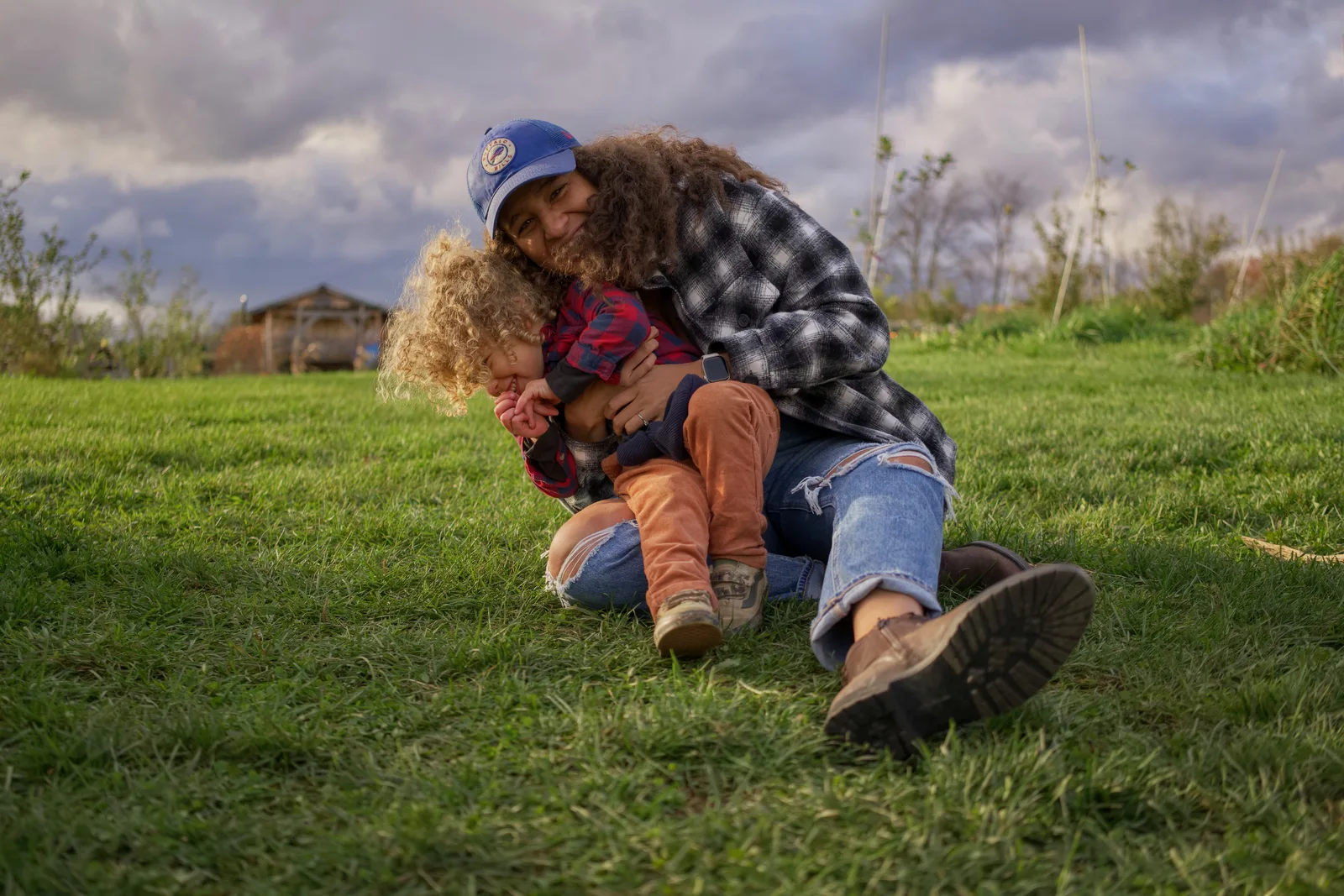 Two people on grassy hill, one in blue cap and plaid shirt embracing child in red plaid shirt