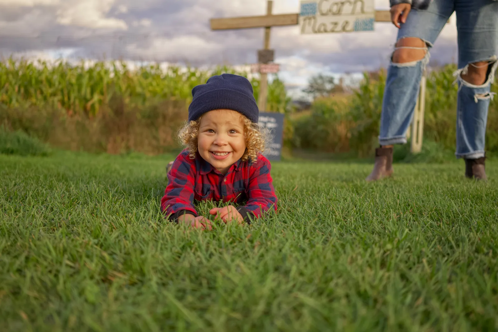 Child in red plaid shirt and dark beanie crawling on grass near Corn Maze sign