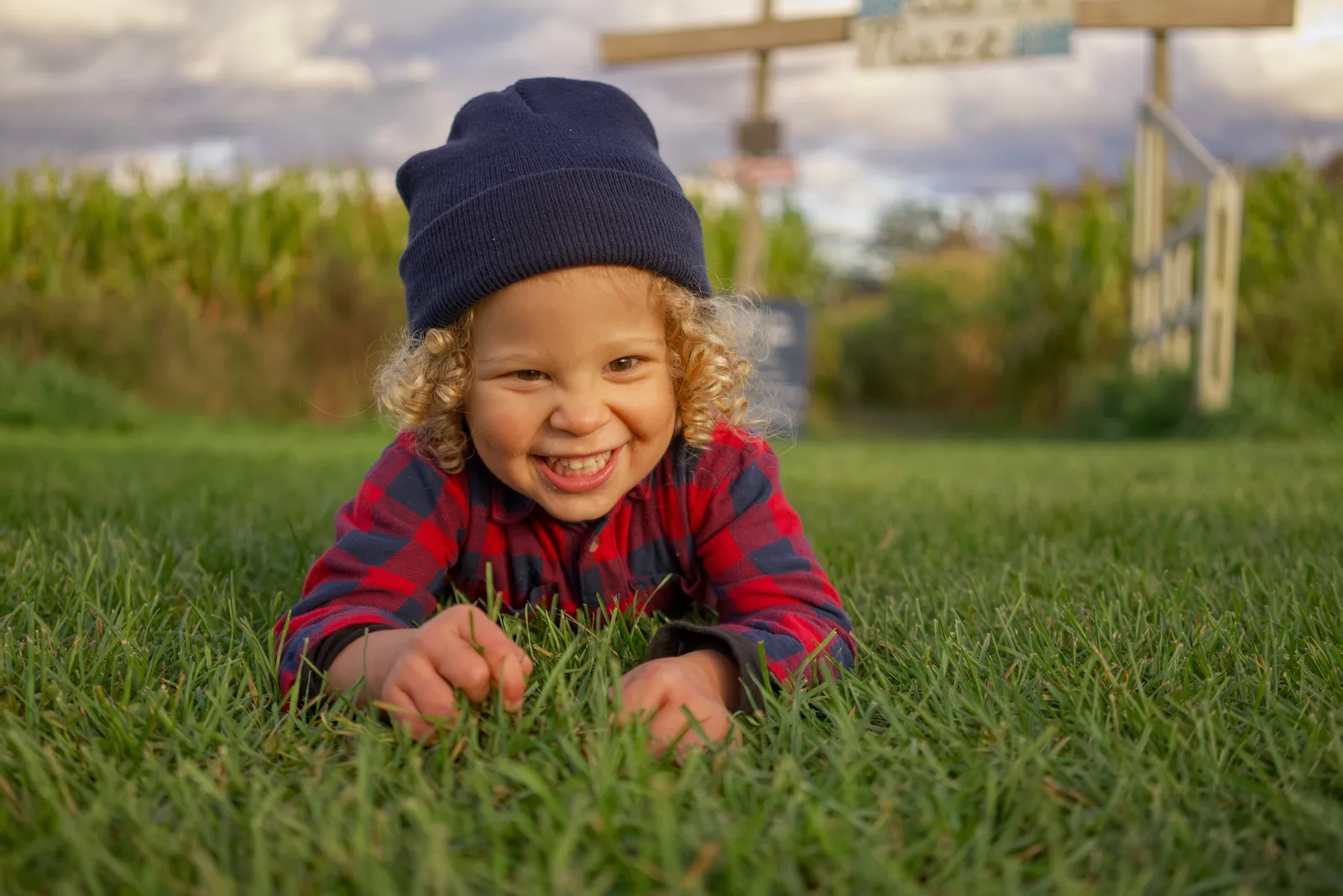 Child in red plaid shirt and blue beanie lying on grass near corn maze
