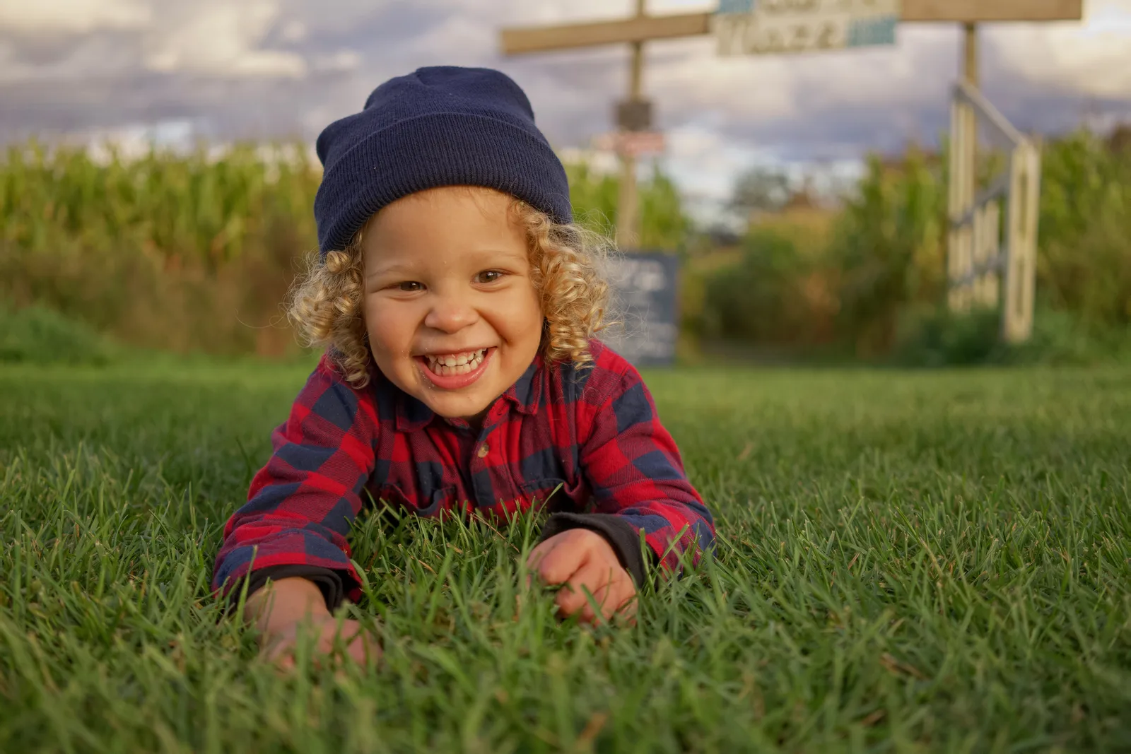 Child in red plaid shirt and blue beanie lying on grass with corn maze sign