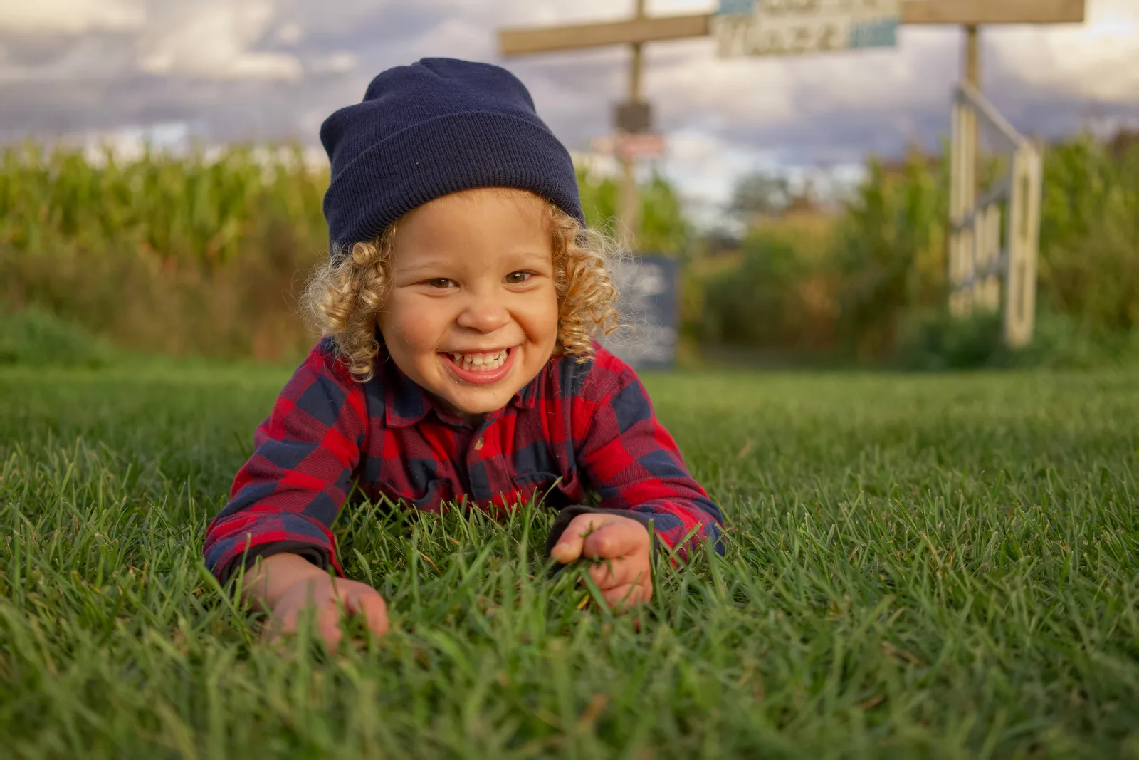 Child in red plaid shirt and blue beanie lying on grass with cloudy sky