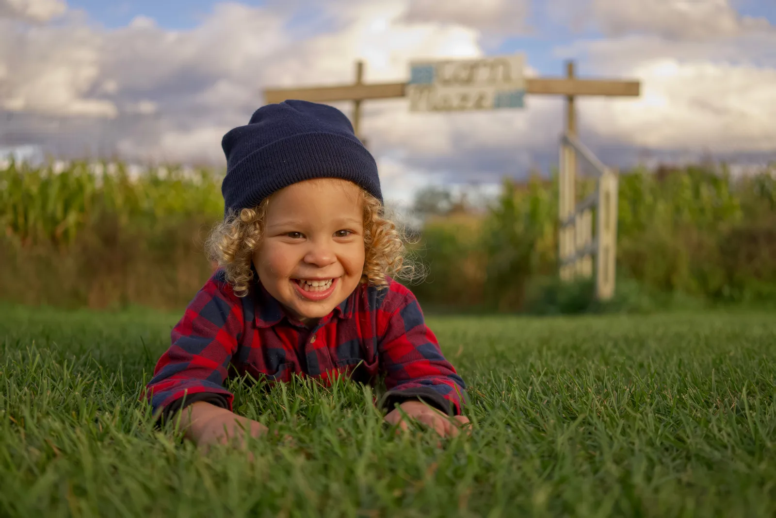 Child in red plaid shirt and blue beanie lying on grass under cloudy sky