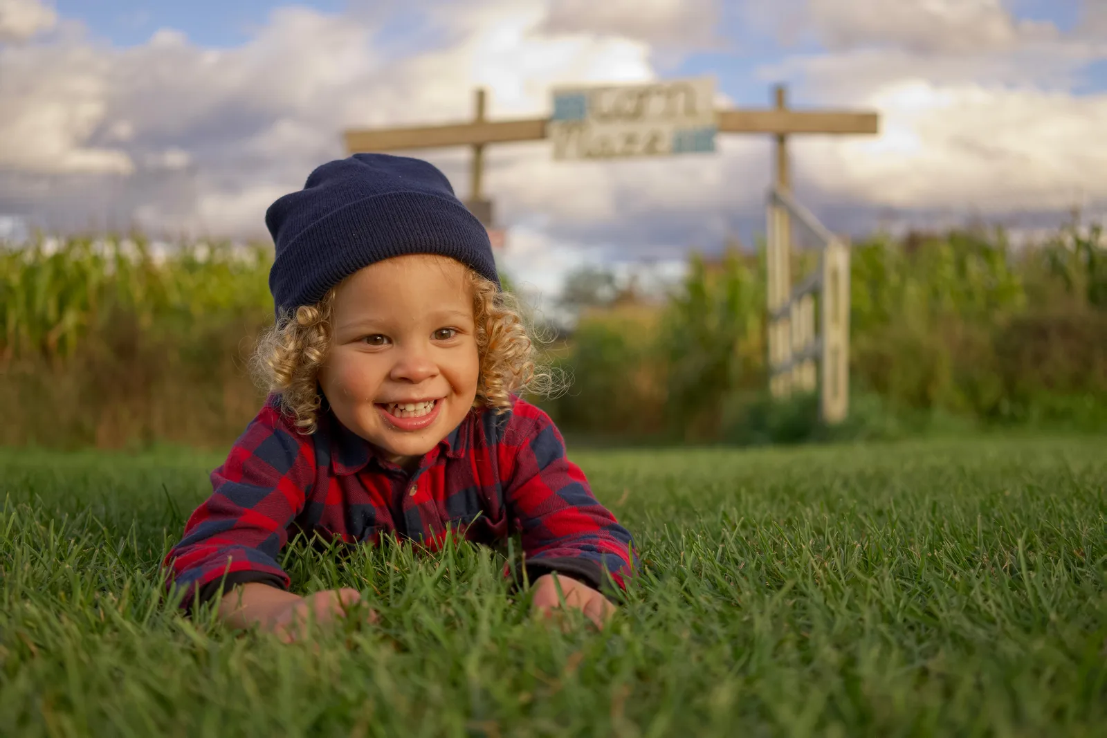 Child in red plaid shirt and blue beanie lying on grass with corn maze sign