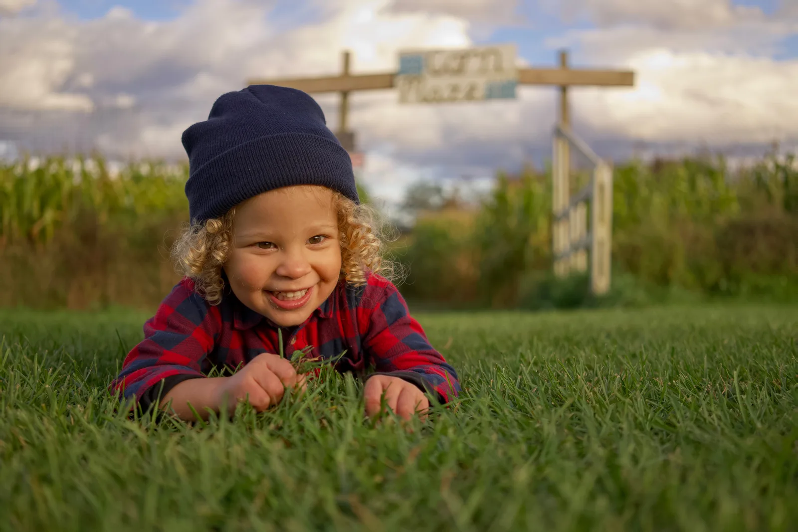 Child in red plaid shirt and blue beanie lying on grass holding grass