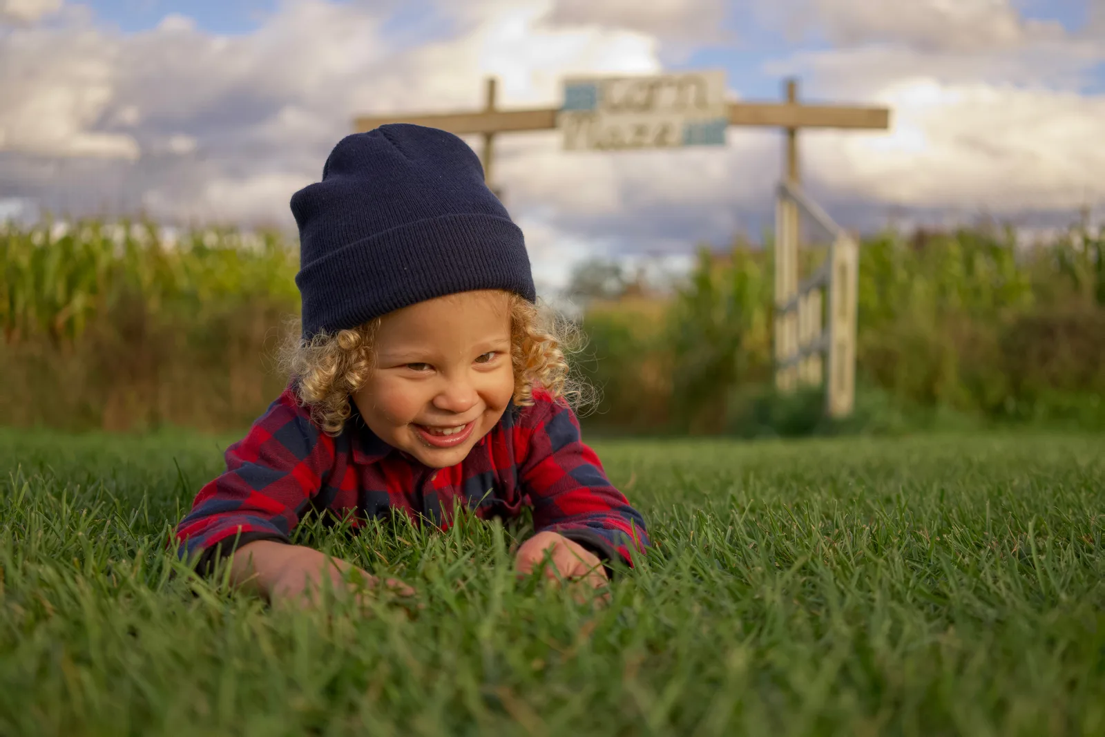 Child in red plaid shirt and dark beanie lying on grass with corn maze sign