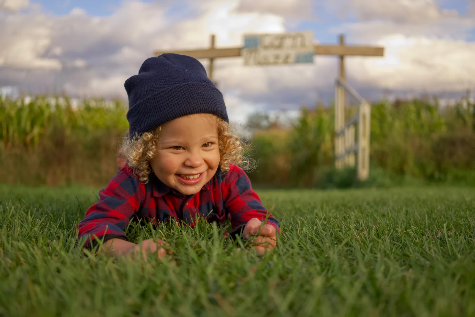 Child in red plaid shirt and dark beanie on grass with corn maze sign and cloudy sky