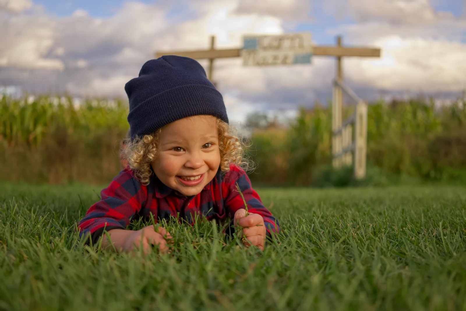 Child in red plaid shirt and dark beanie on grass with corn maze sign and fence