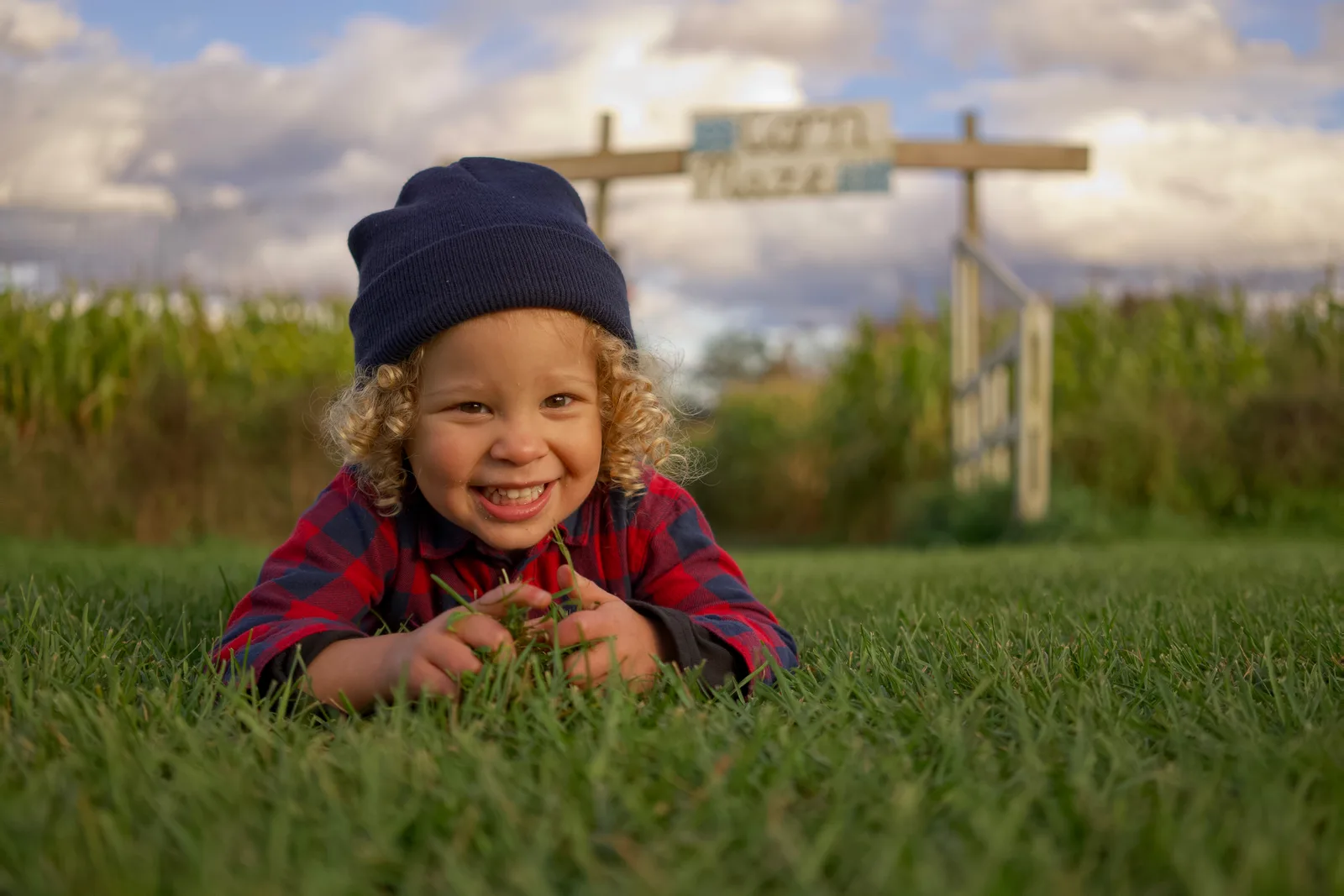 Child in red plaid shirt and dark beanie on grass with corn maze sign and fence
