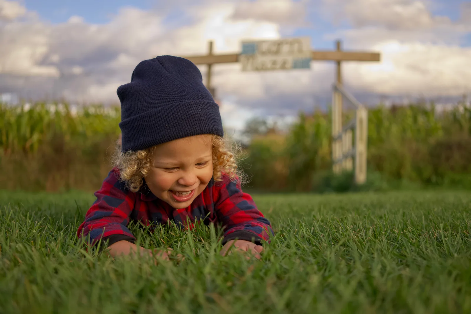 Child in red plaid shirt and dark beanie on grass with corn maze sign and cloudy sky