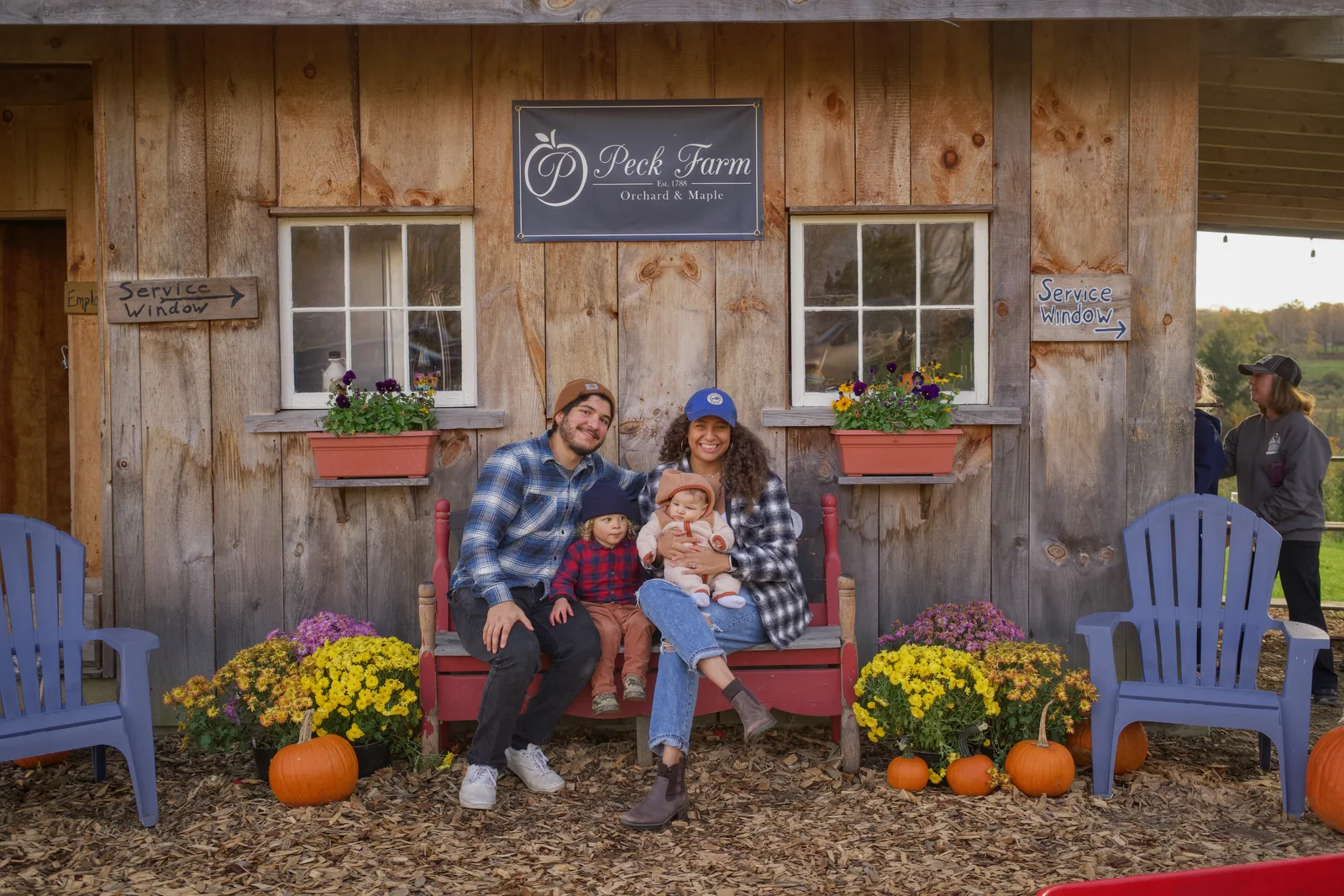 Four people on red bench by wooden building with flowers, pumpkins, and blue chairs