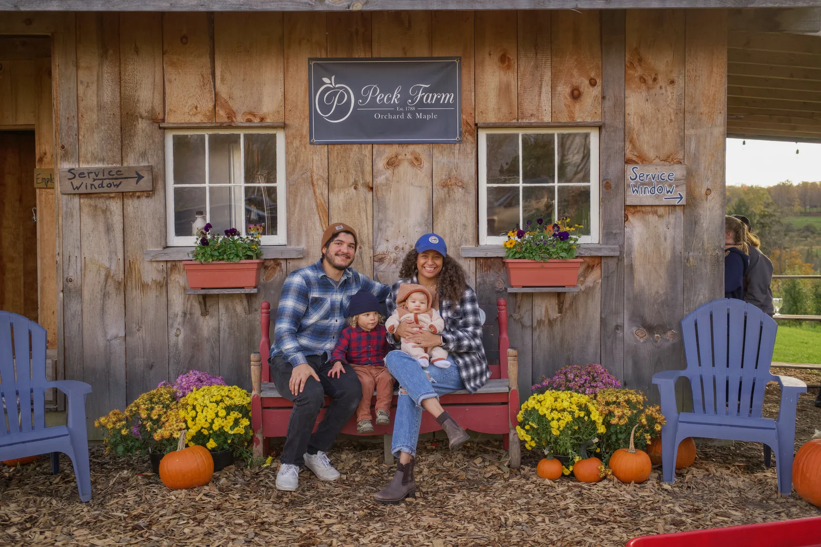 Four people on red bench by wooden building, pumpkins and flowers in foreground