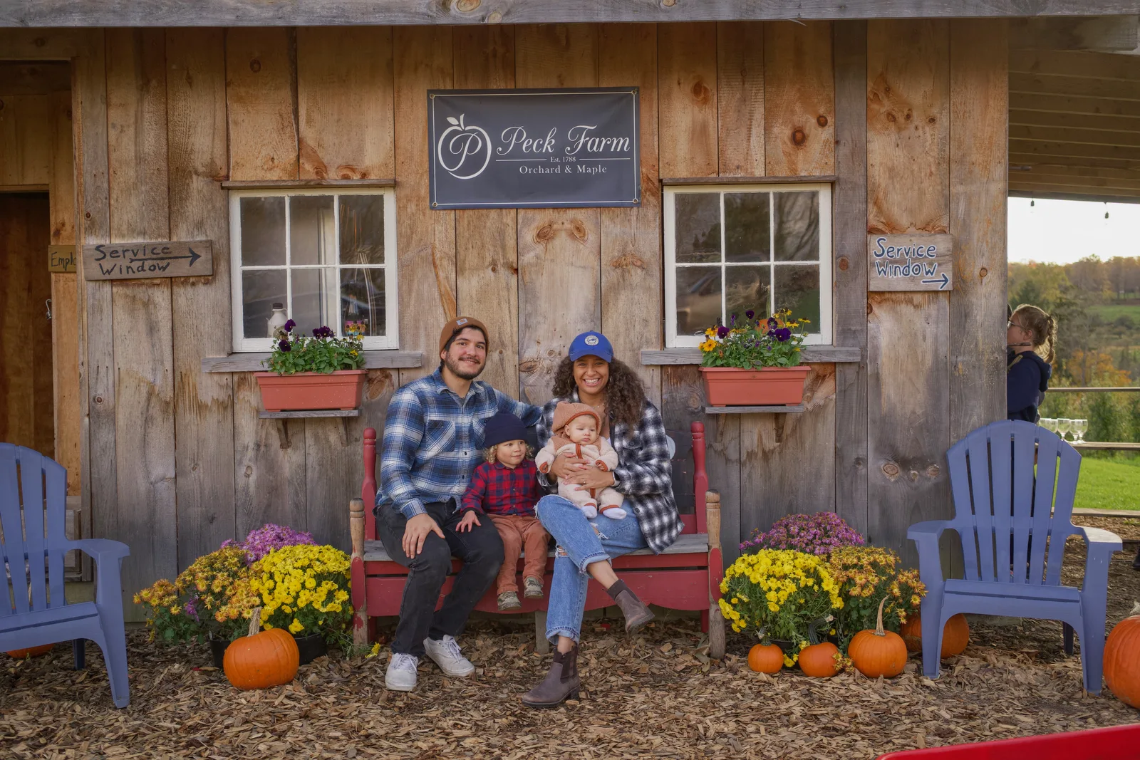Four people on red bench by wooden building, pumpkins and flowers at their feet