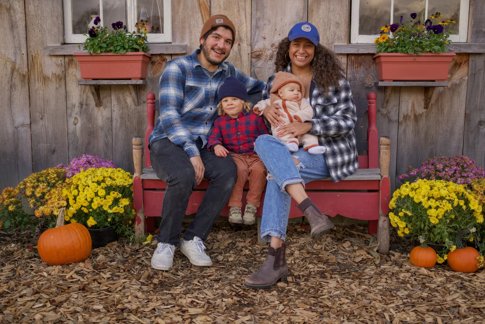 Four people on red bench by wooden building, pumpkins and yellow flowers nearby