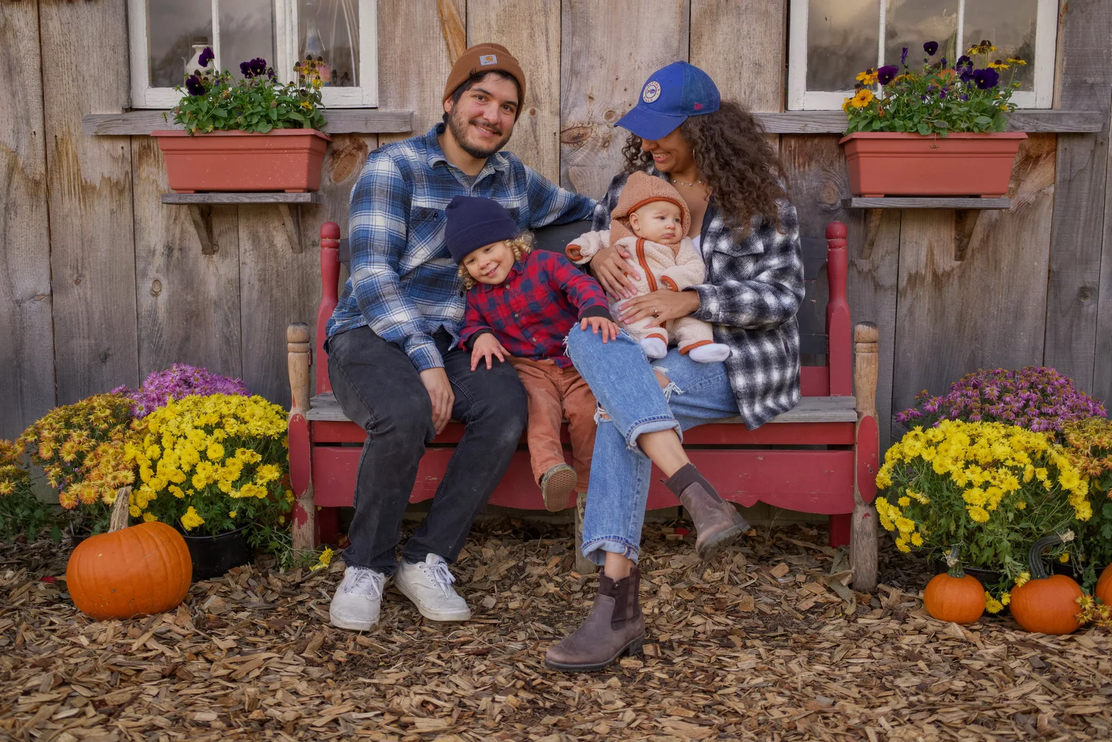 Four people on red bench by wooden building, pumpkins and purple flowers around