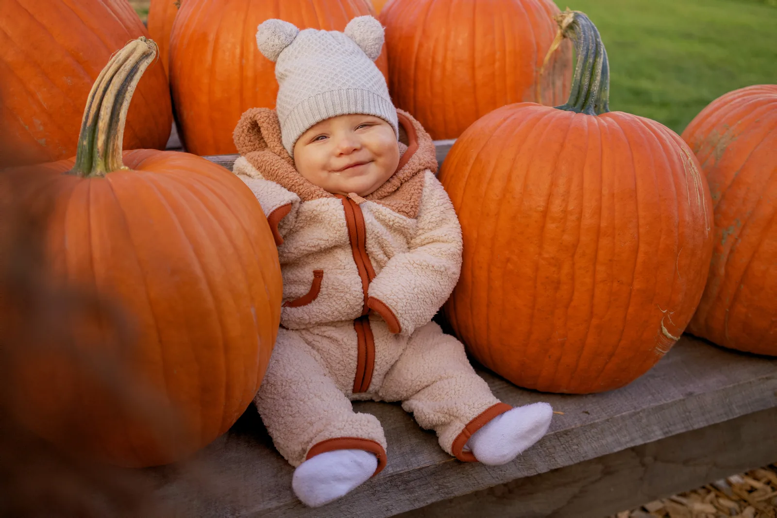 Child in white beanie and bear-hooded onesie sitting on wooden platform surrounded by large orange pumpkins