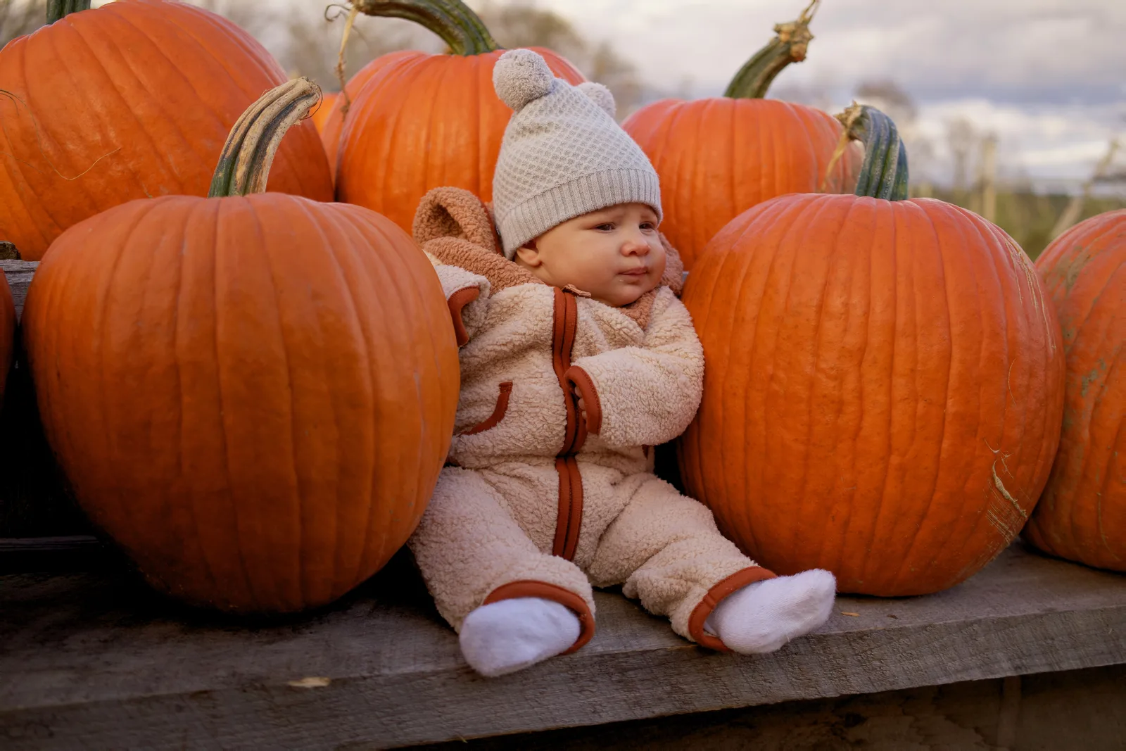 Child in white beanie and bear-hooded onesie sitting on wooden platform surrounded by large orange pumpkins
