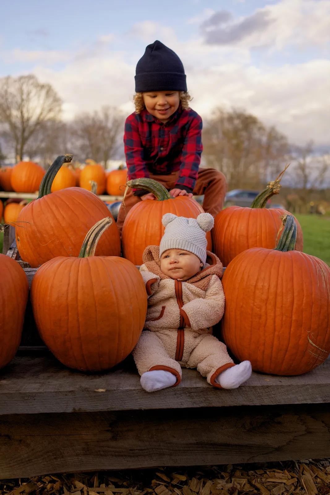 Two children on wooden platform with large orange pumpkins, one in plaid shirt and navy beanie crouching