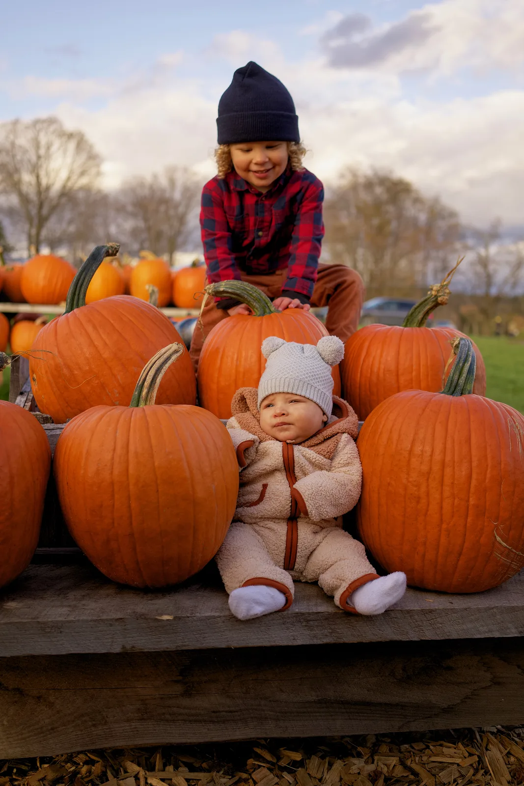 Two children on wooden platform with large orange pumpkins, one in plaid shirt and navy beanie kneeling