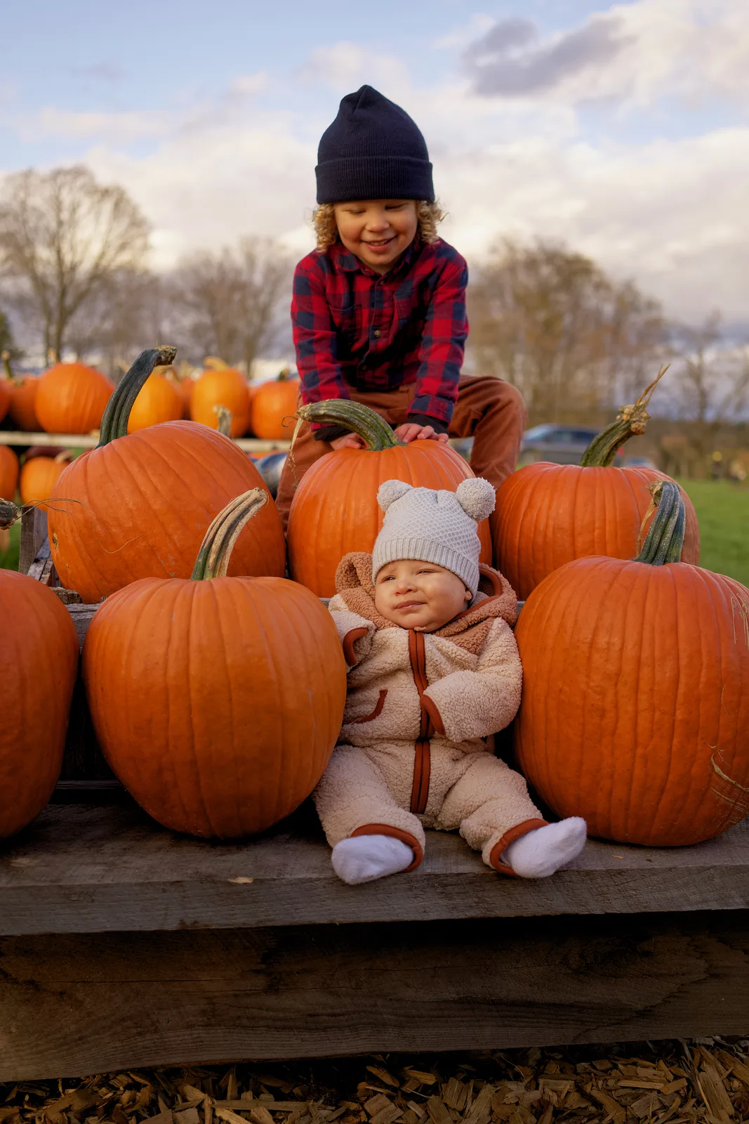 Two children on wooden platform with large orange pumpkins, one in plaid shirt and navy beanie leaning forward