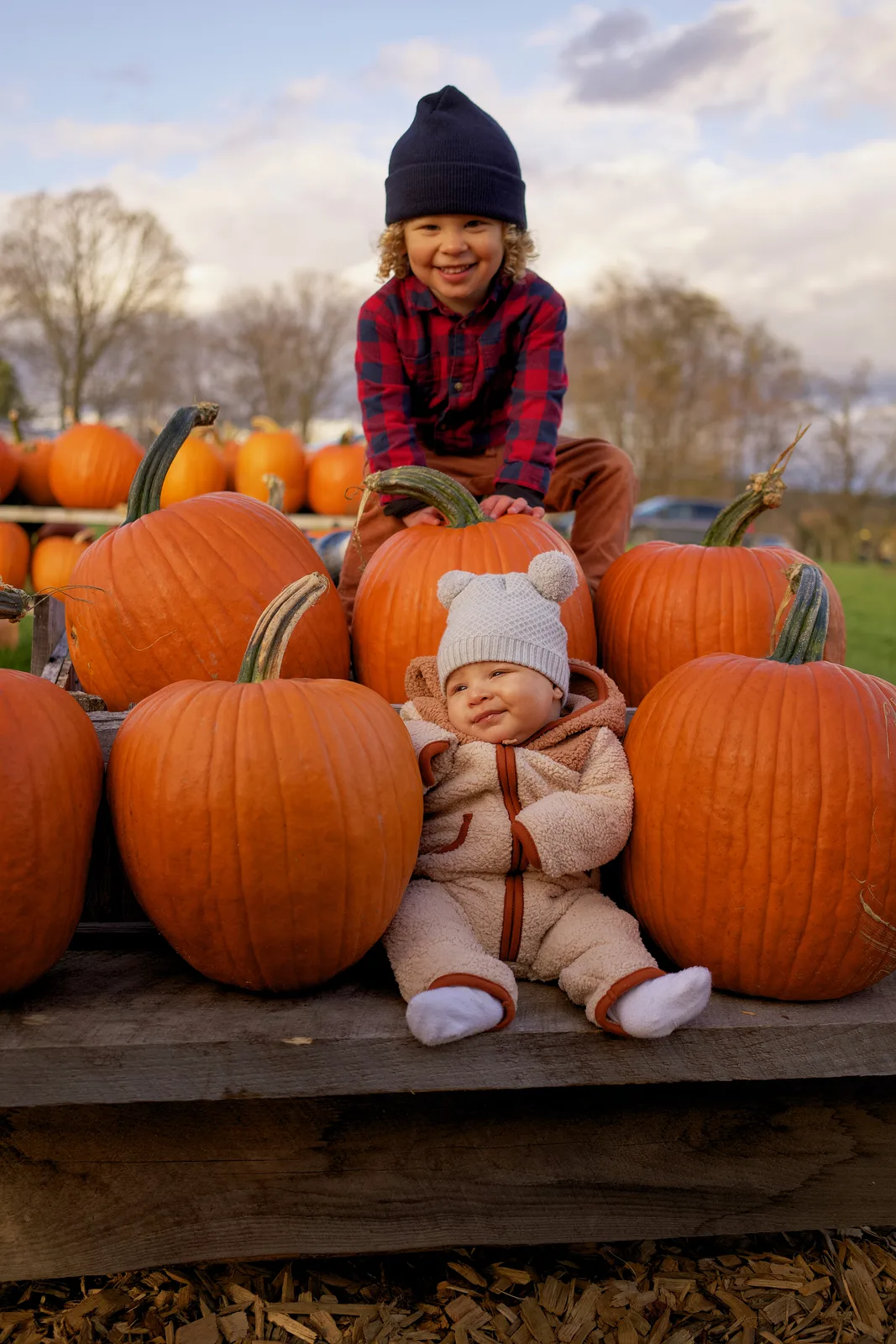 Two children on wooden platform beside large orange pumpkins, one in plaid shirt and navy beanie