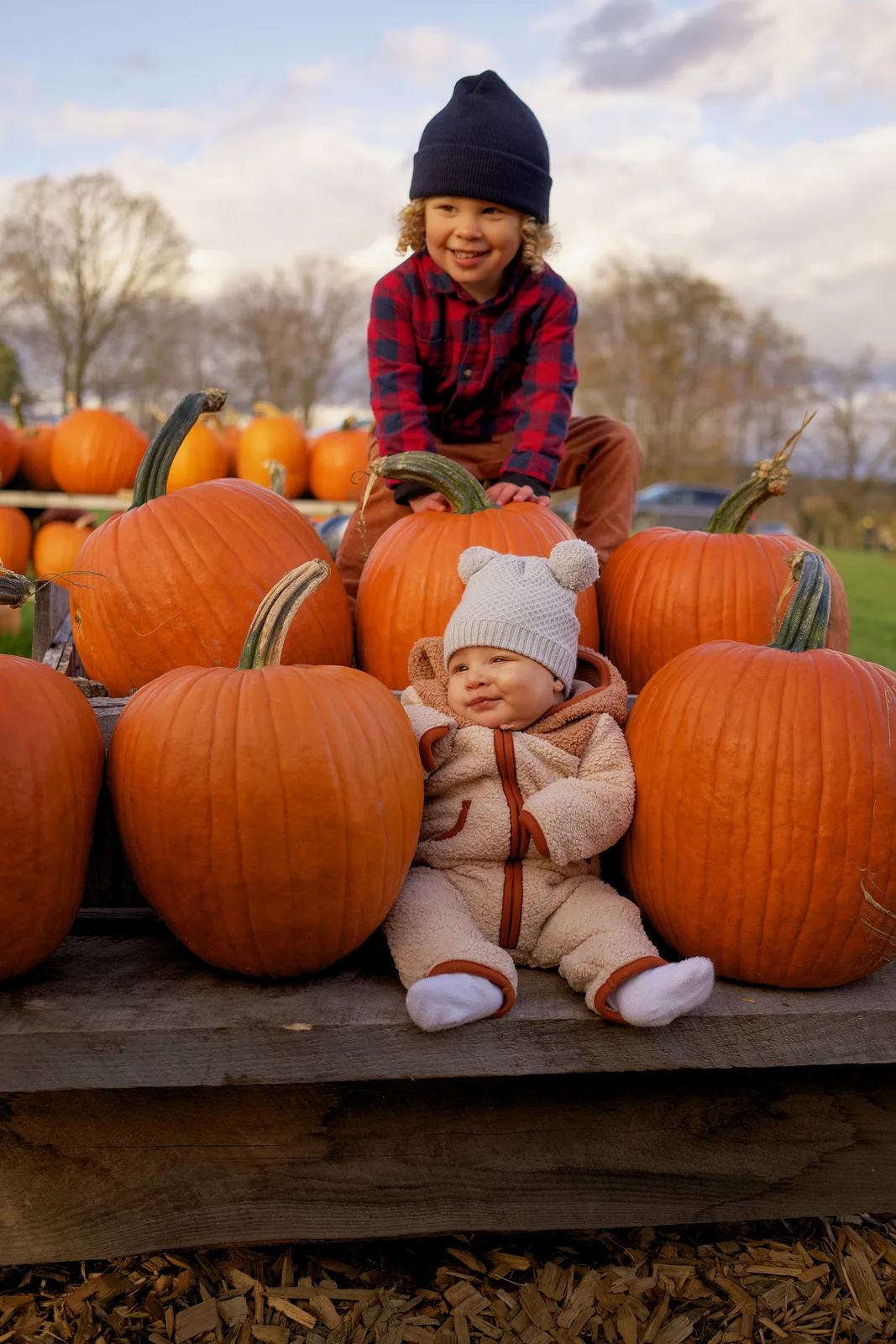 Two children on wooden platform with large orange pumpkins, one in plaid shirt and navy beanie smiling