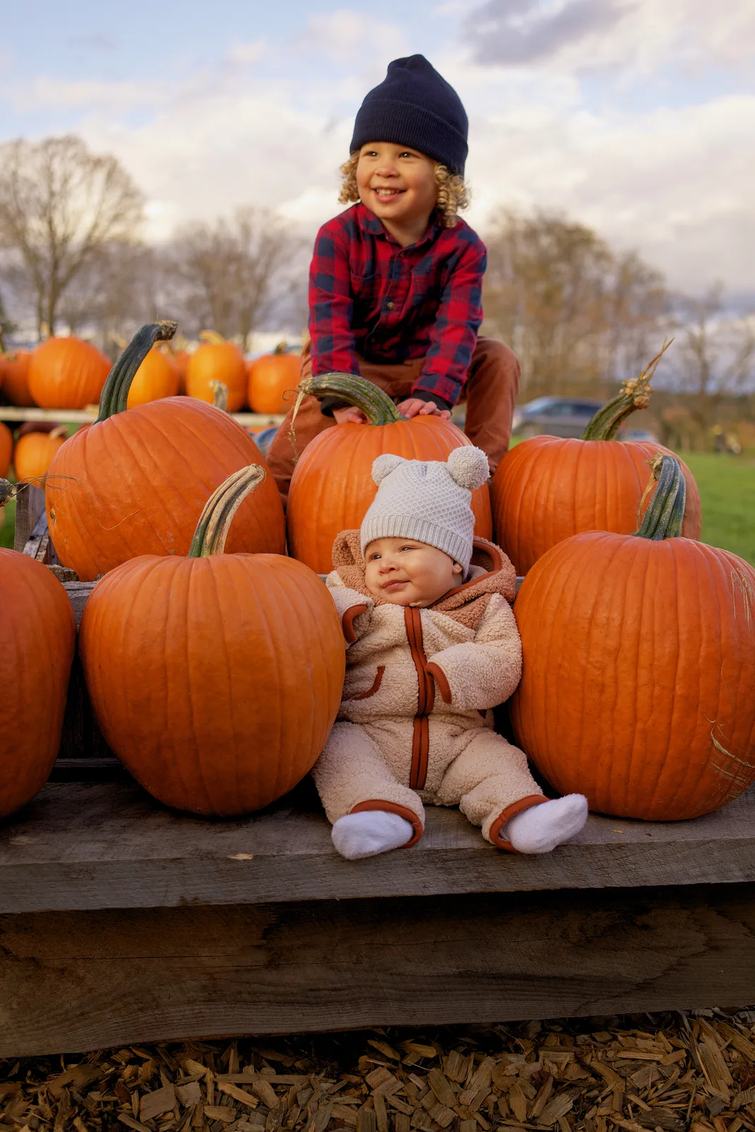 Two children on wooden platform surrounded by large orange pumpkins, one in plaid shirt and navy beanie