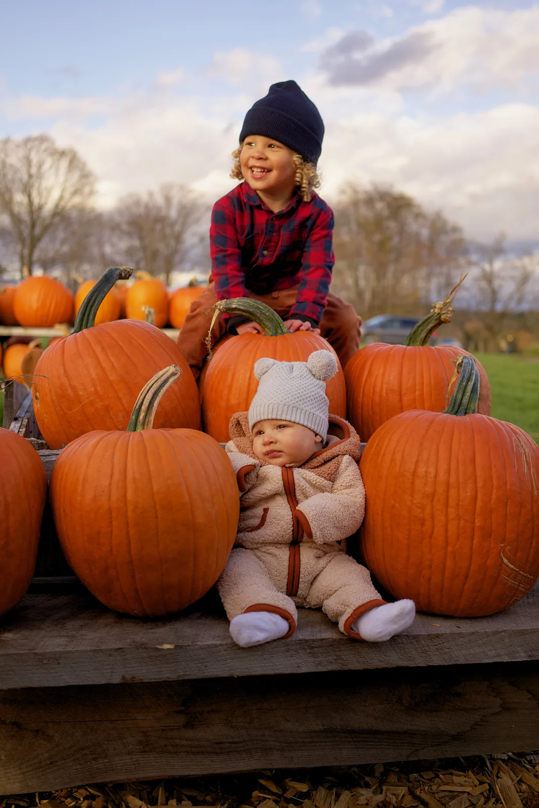 Two children on wooden platform with large orange pumpkins, one in plaid shirt and navy beanie smiling