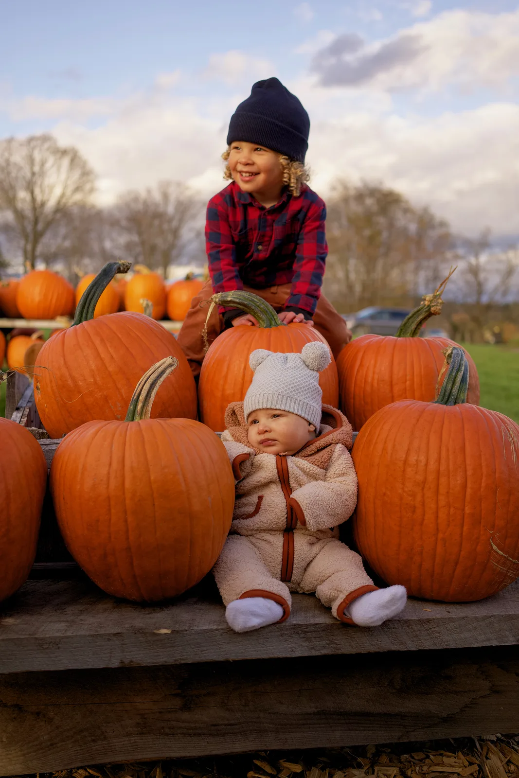 Two children on wooden platform beside large orange pumpkins, one in plaid shirt and navy beanie
