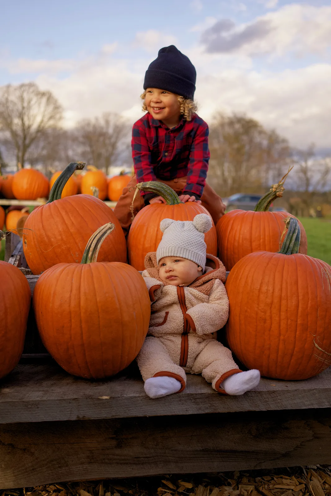 Two children on wooden platform with large orange pumpkins around, one in plaid shirt and navy beanie