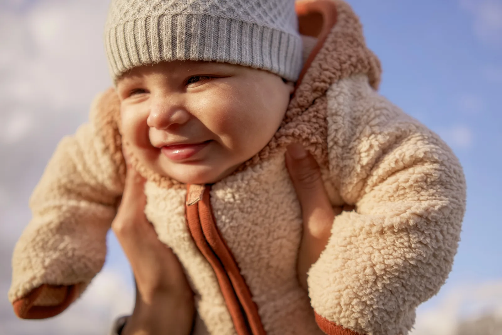 Child in light gray knit hat and cream bear-hooded onesie with hands near face