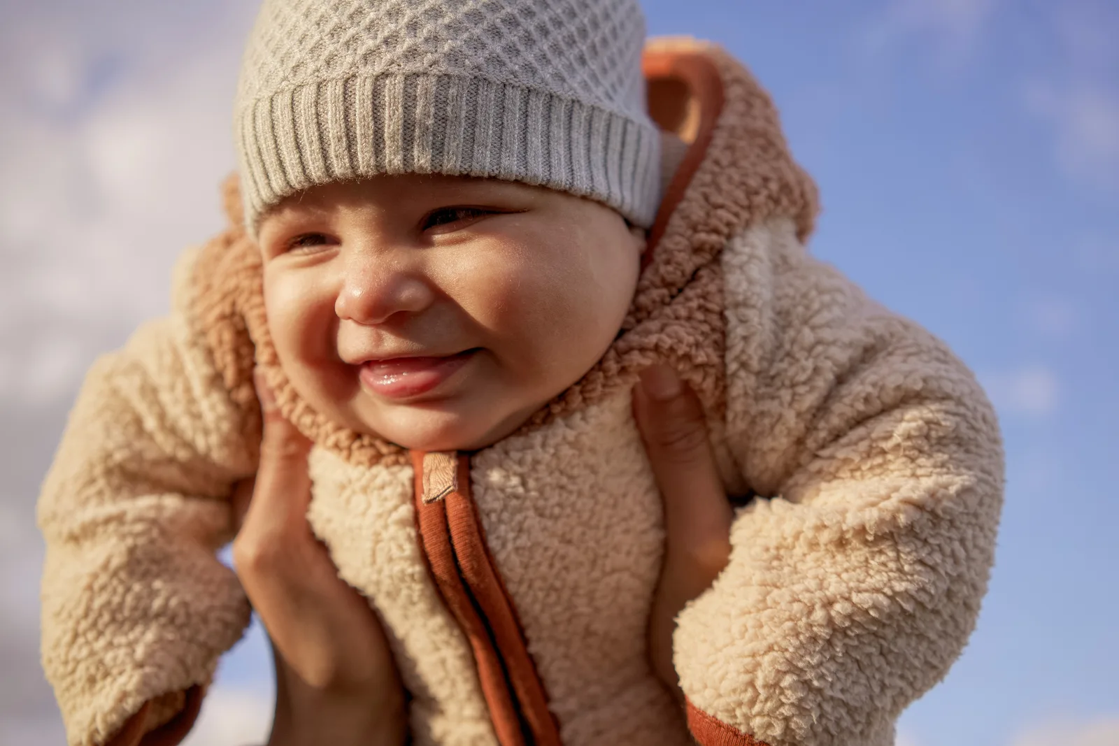 Child in light gray knit hat and cream bear-hooded onesie smiling with hands near face