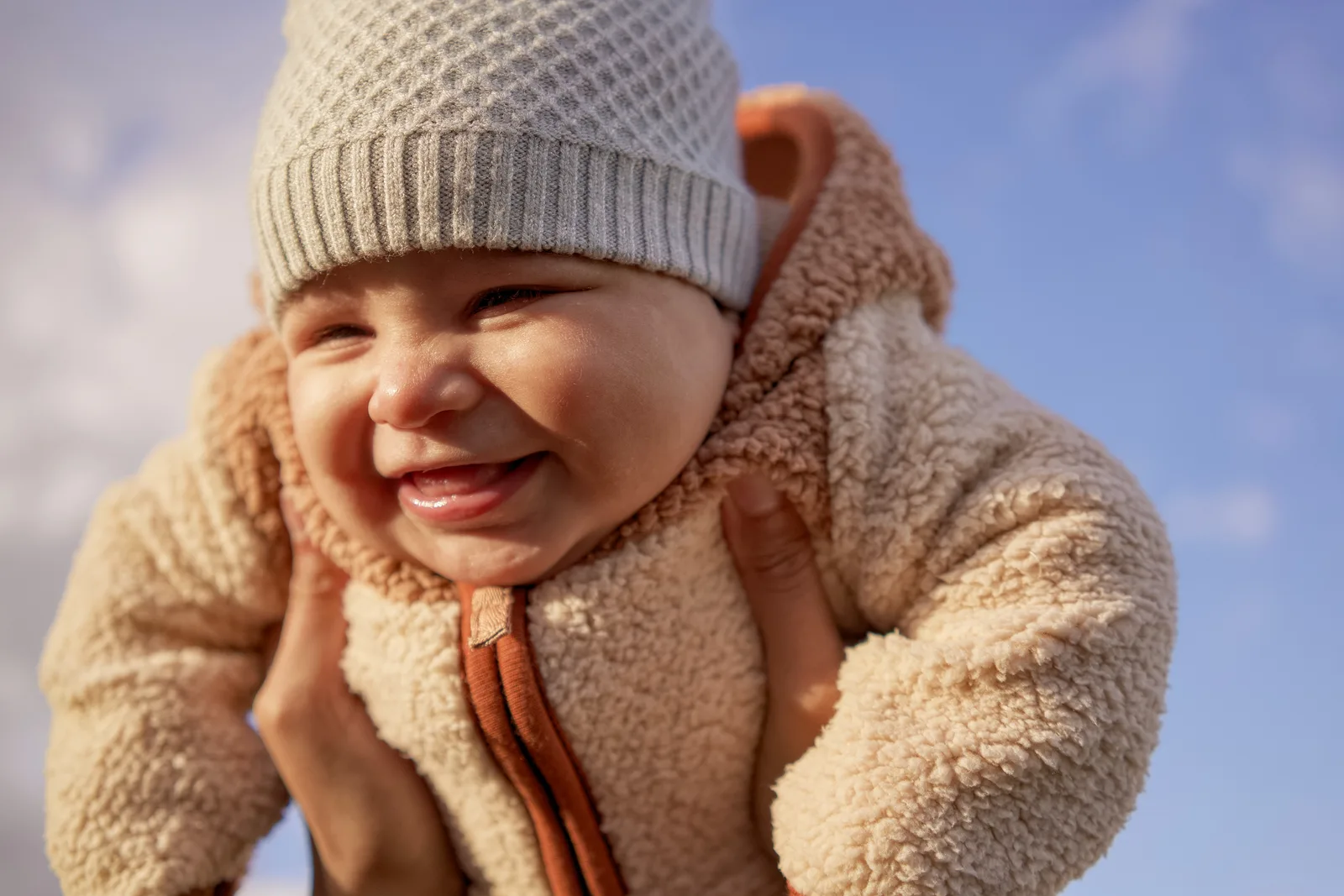Child in light gray knit hat and cream bear-hooded onesie laughing with hands near face