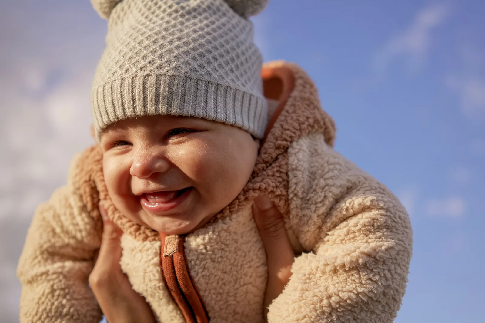 Child in light gray knit hat and cream bear-hooded onesie with hands near face