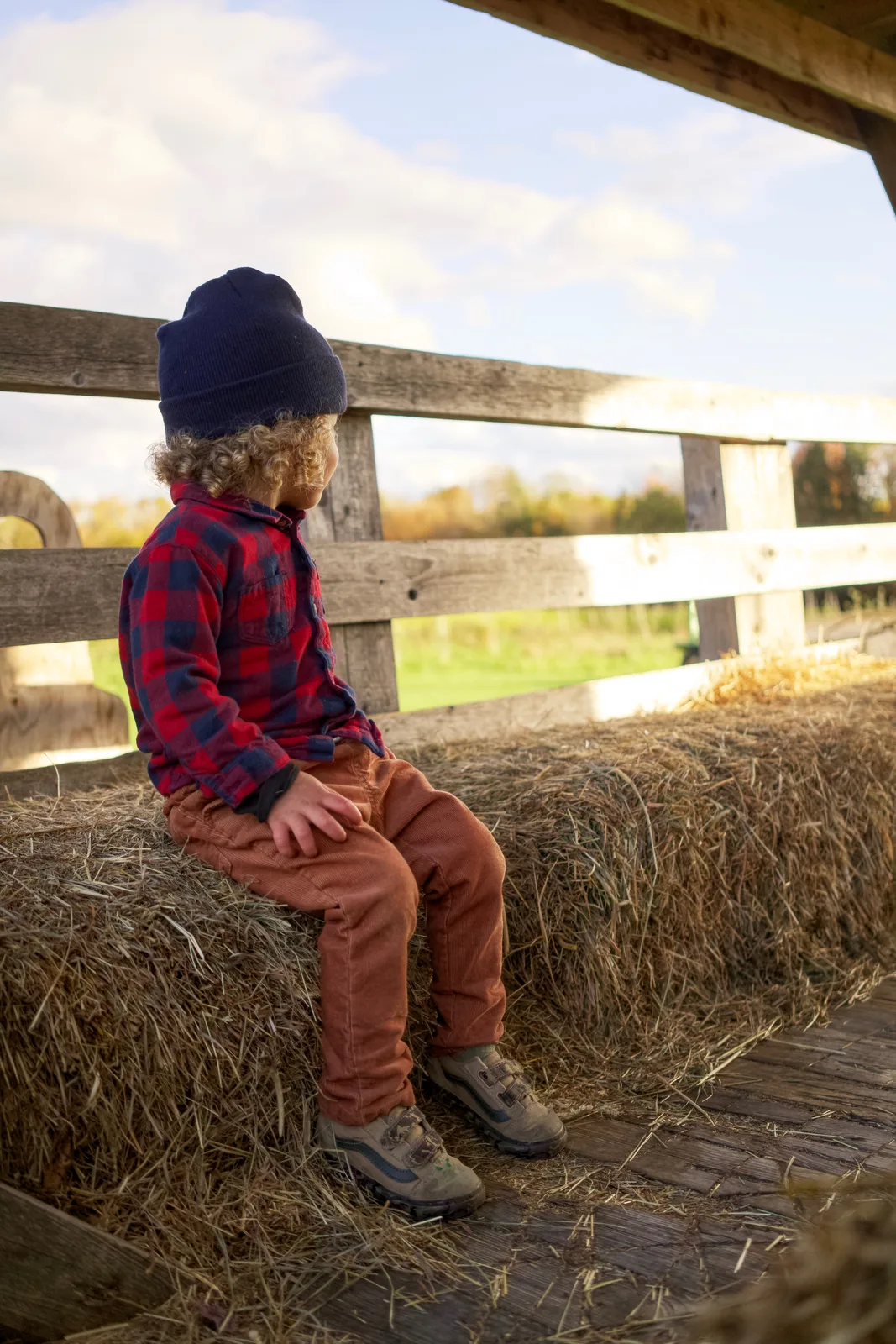 Child in red plaid shirt and navy beanie sitting on hay bale by wooden fence