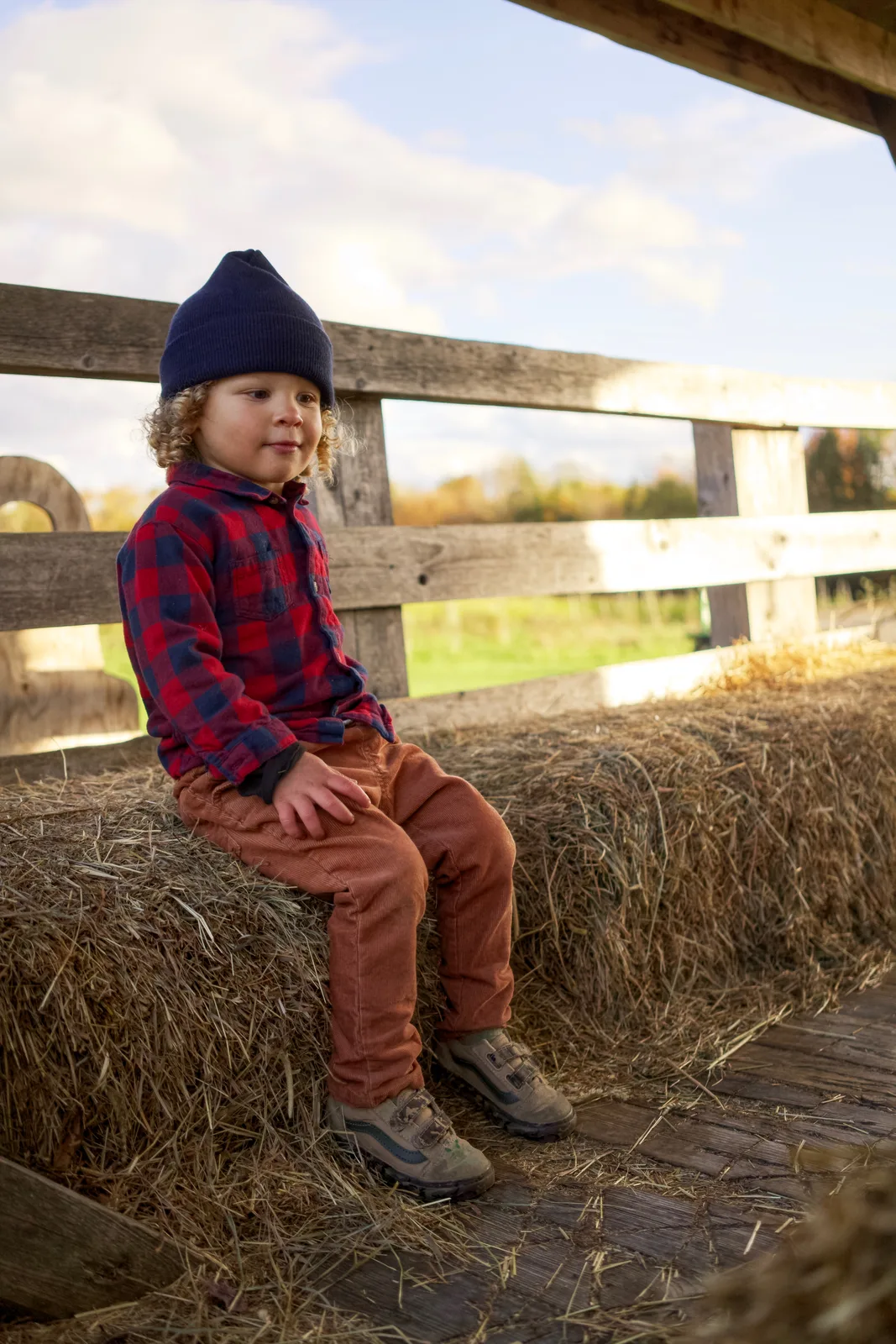 Child in red plaid shirt and navy beanie sitting on hay bale, hands on knees