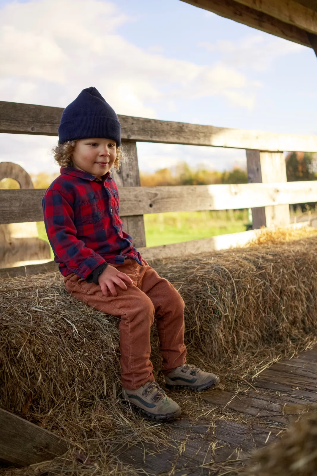 Child in red plaid shirt and navy beanie sitting on hay bale, looking to the side