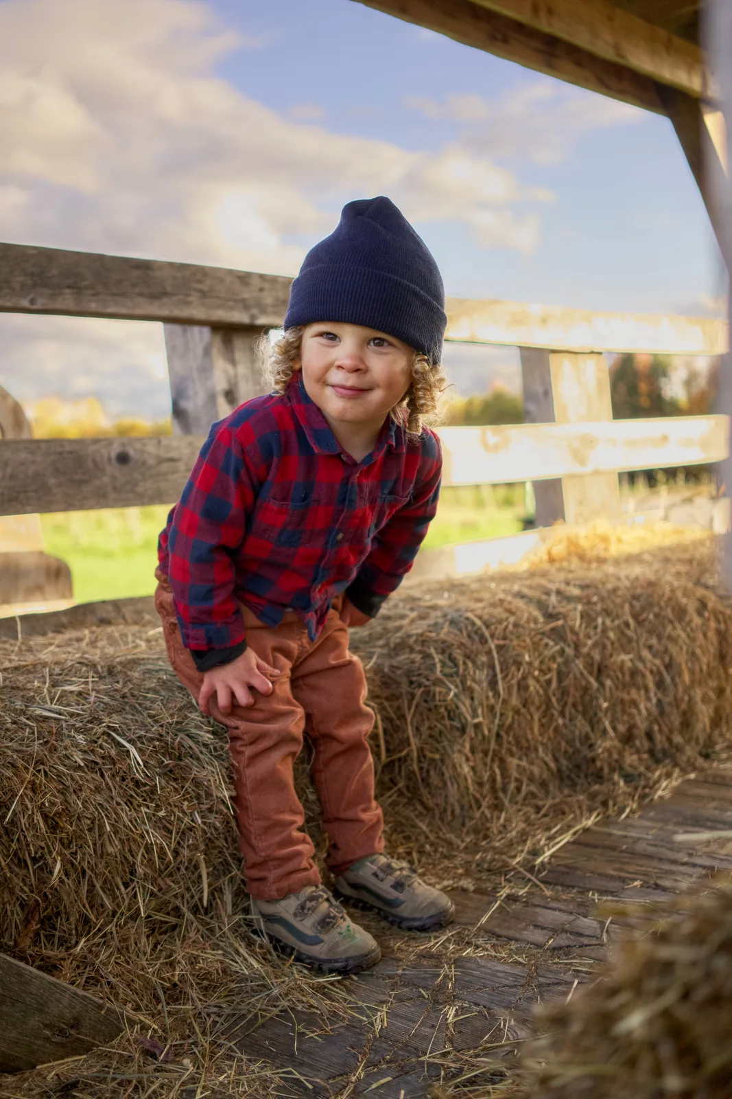 Child in navy beanie, red plaid shirt, and brown pants standing on hay bale