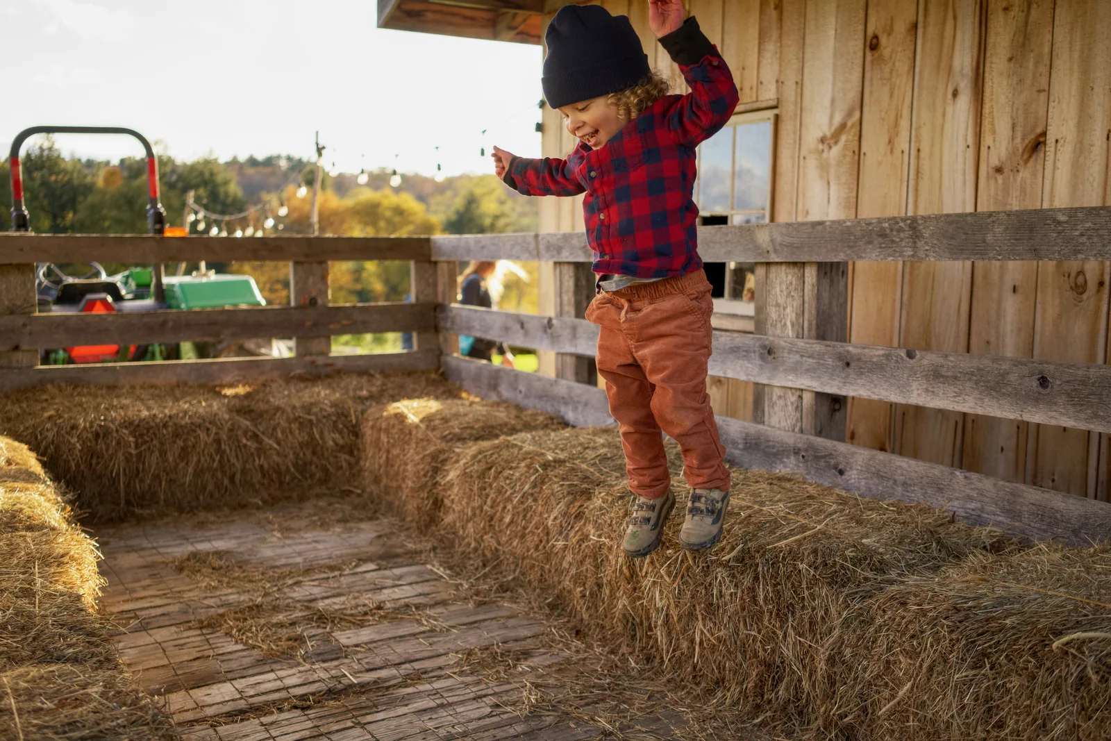 Child in navy beanie, red plaid shirt, and brown pants jumping on hay bales