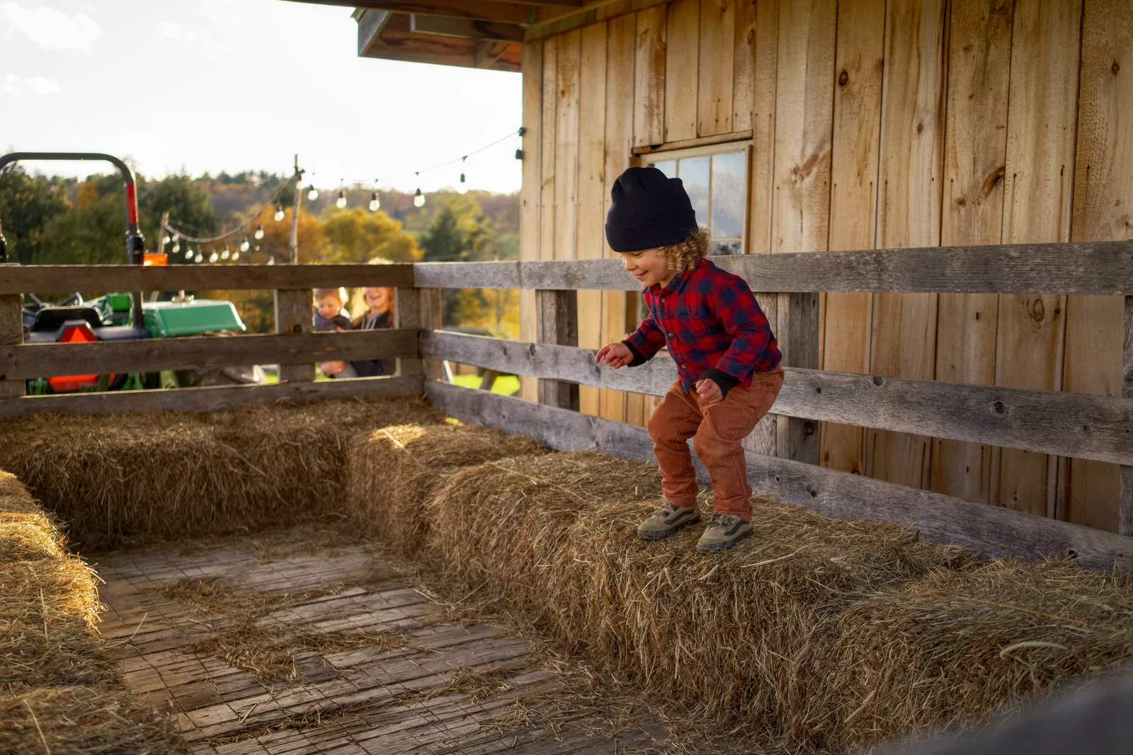 Child in navy beanie, red plaid shirt, and brown pants stepping on hay bales