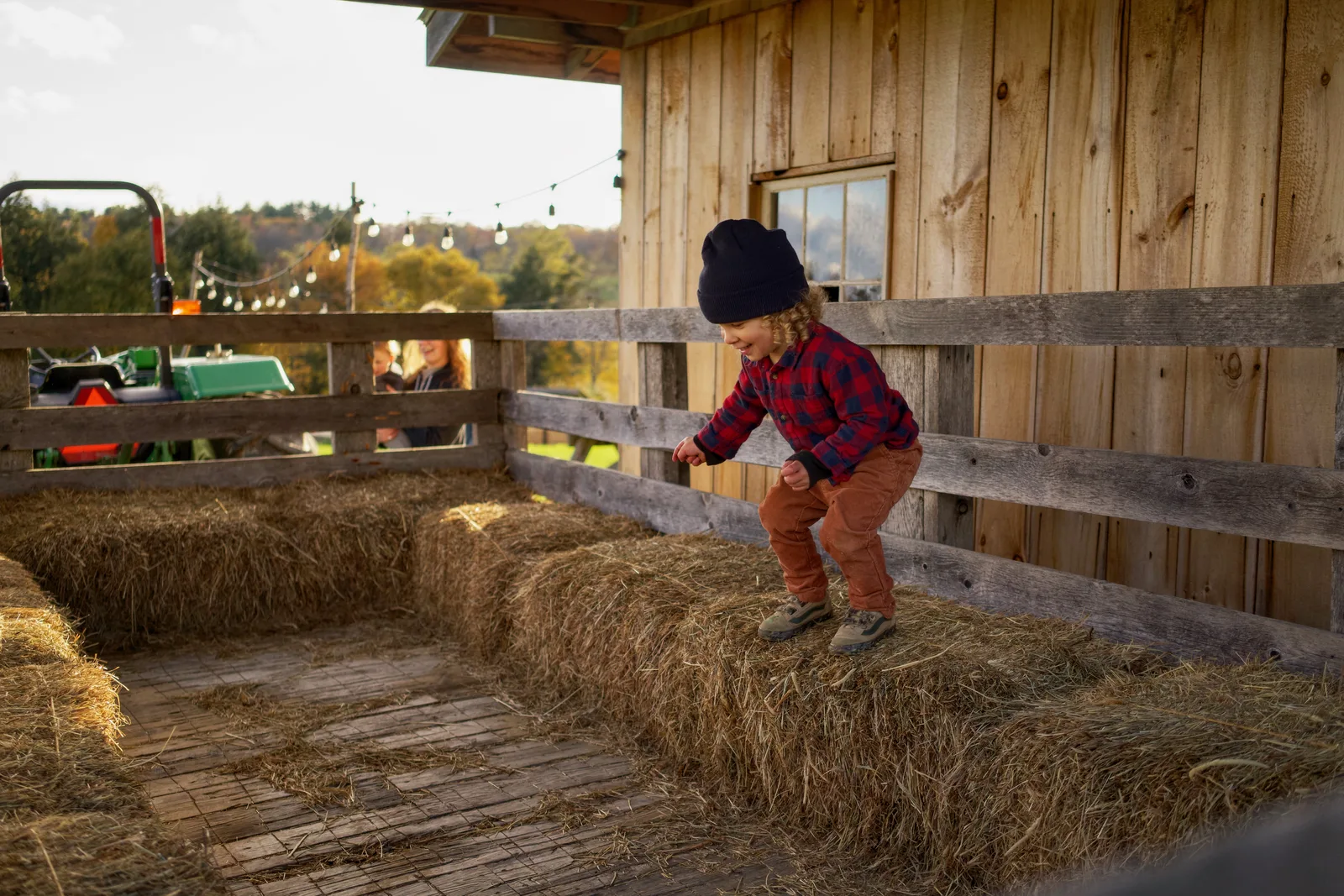 Child in black beanie, red plaid shirt, and orange pants jumping on hay bales in barn