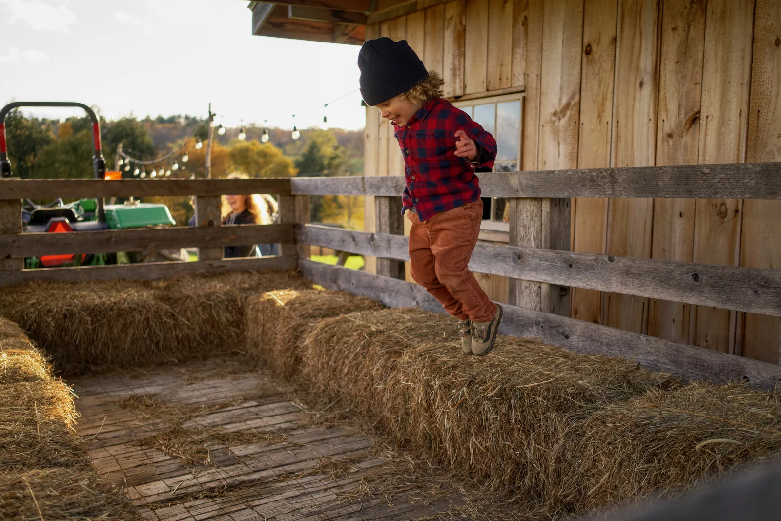 Child in black beanie, red plaid shirt, and orange pants mid-jump on hay bales